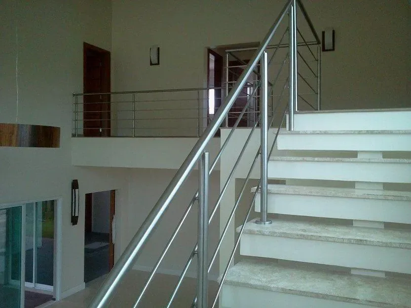 Guarda-corpo em inox com tubos horizontais instalado em escada de mármore de residência ampla, com mezanino ao fundo.