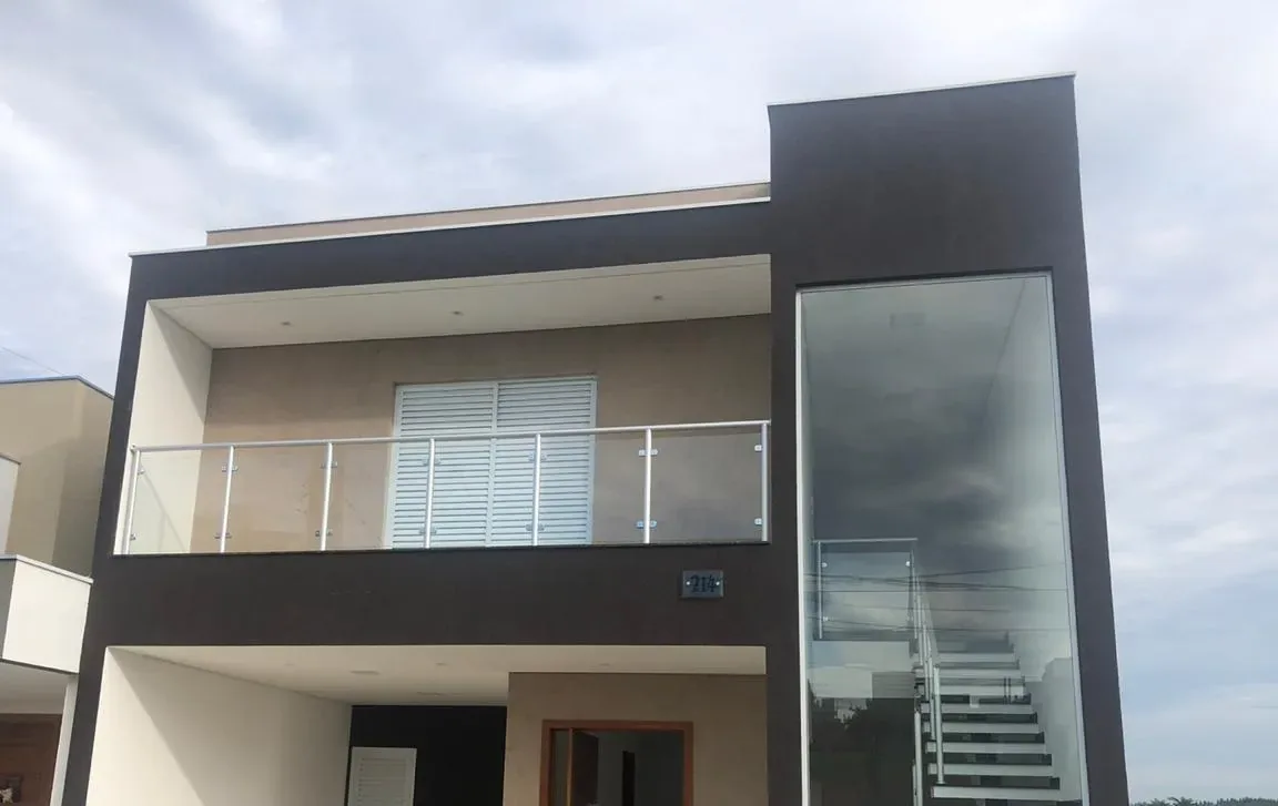 Fachada de casa moderna com guarda-corpo misto de tubo em alumínio branco e vidro temperado na sacada do segundo pavimento, ao lado de janela de vidro estrutural do piso ao teto.