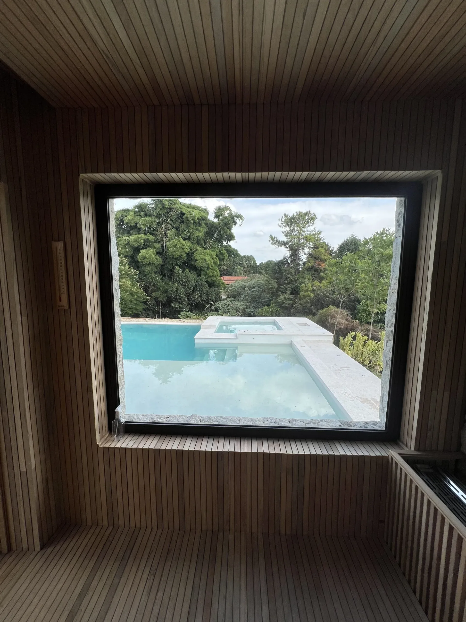 Janela fixa em alumínio preto emoldurada por revestimento de madeira de sauna, com vista panorâmica para piscina e jardim arborizado.