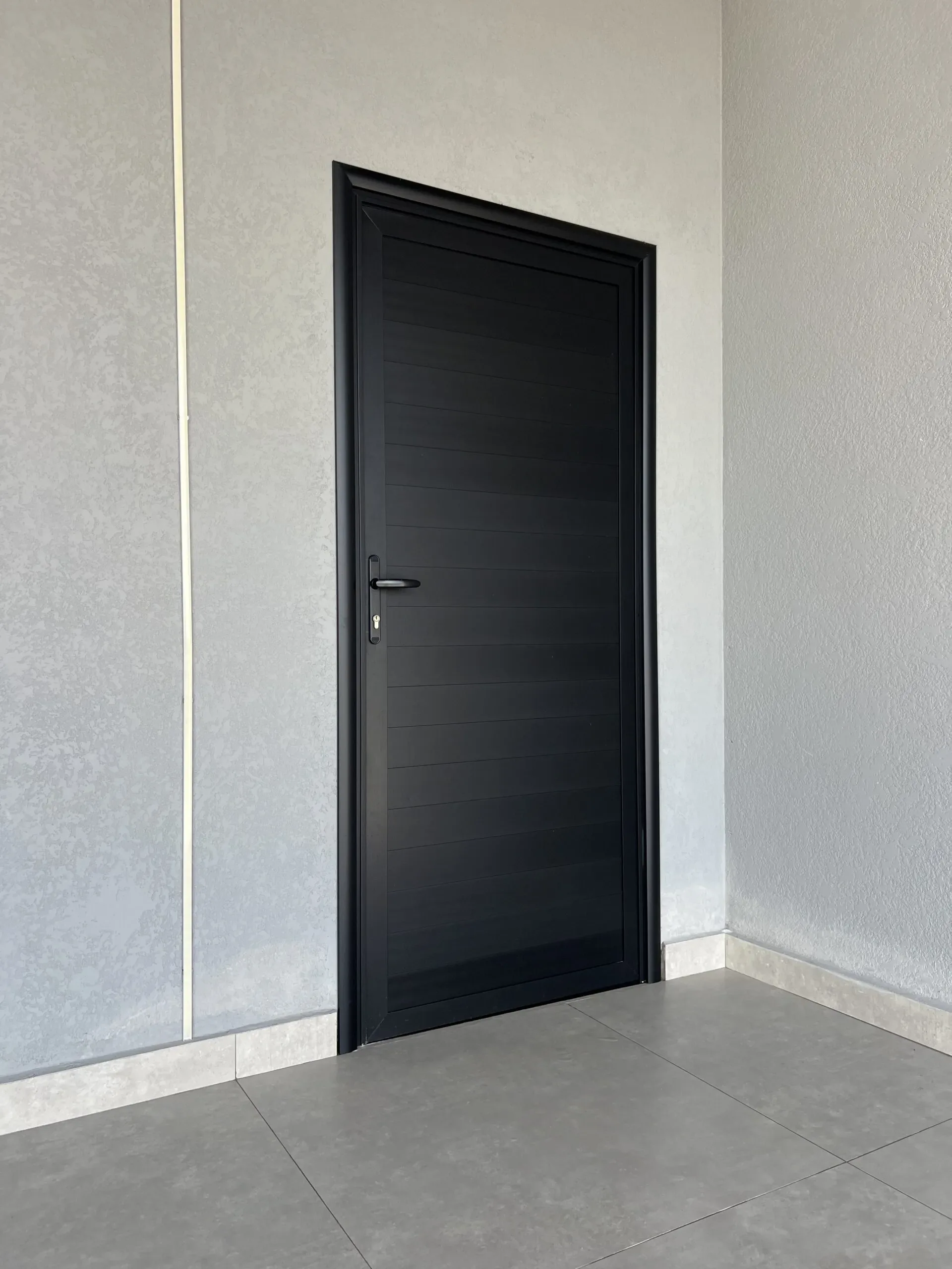 Porta de abrir em alumínio preto com textura de ripas horizontais, sem vidro, instalada em parede rústica.