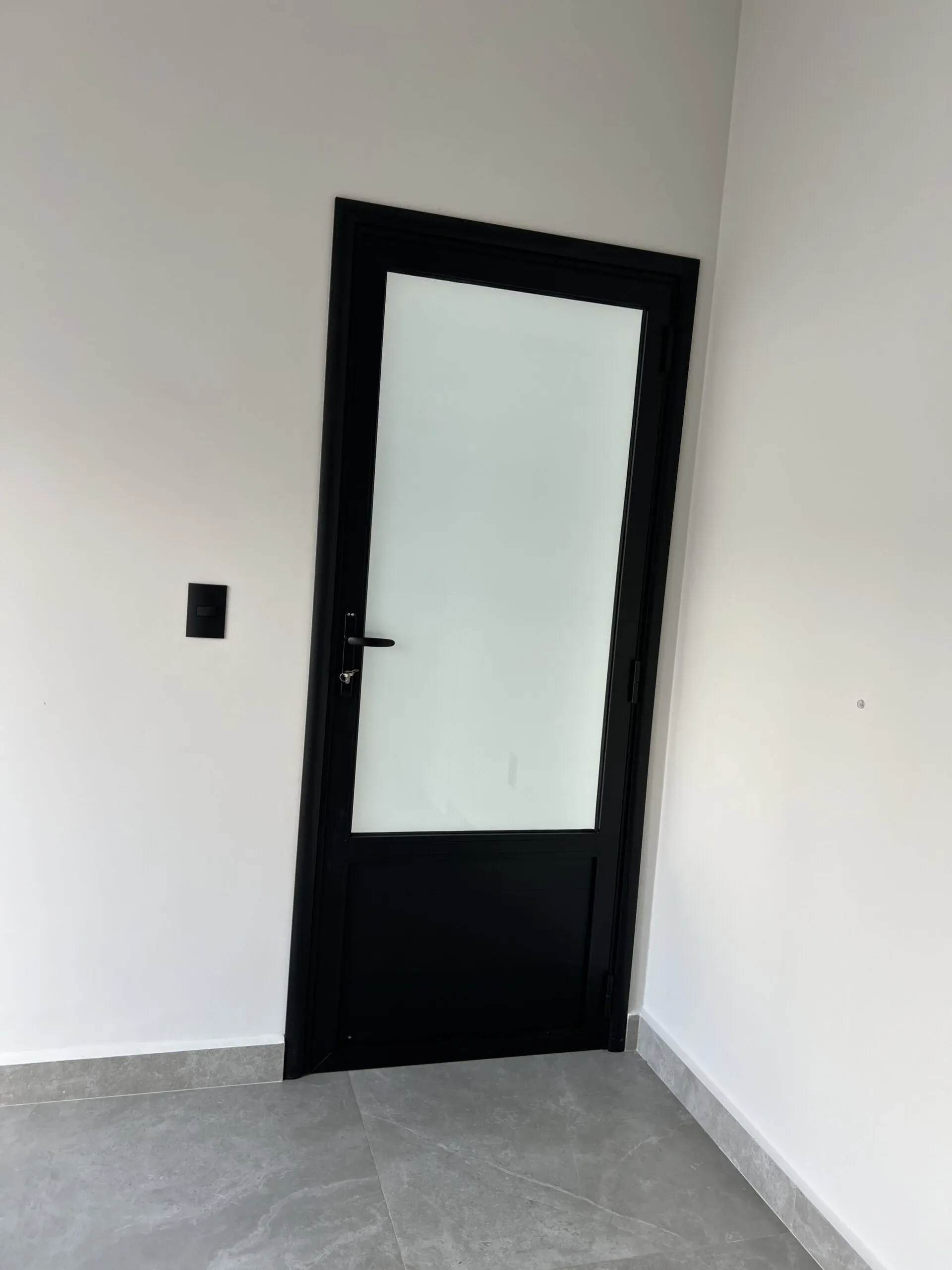 Porta de abrir de alumínio preto com painel de vidro leitoso na parte superior sobre piso cinza.