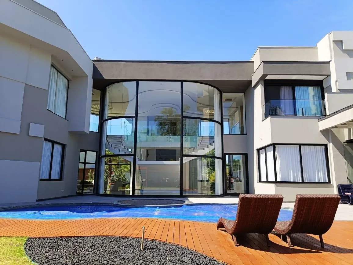 Fachada curva de vidro com esquadrias de alumínio pretas em casa de alto padrão com piscina e deck de madeira