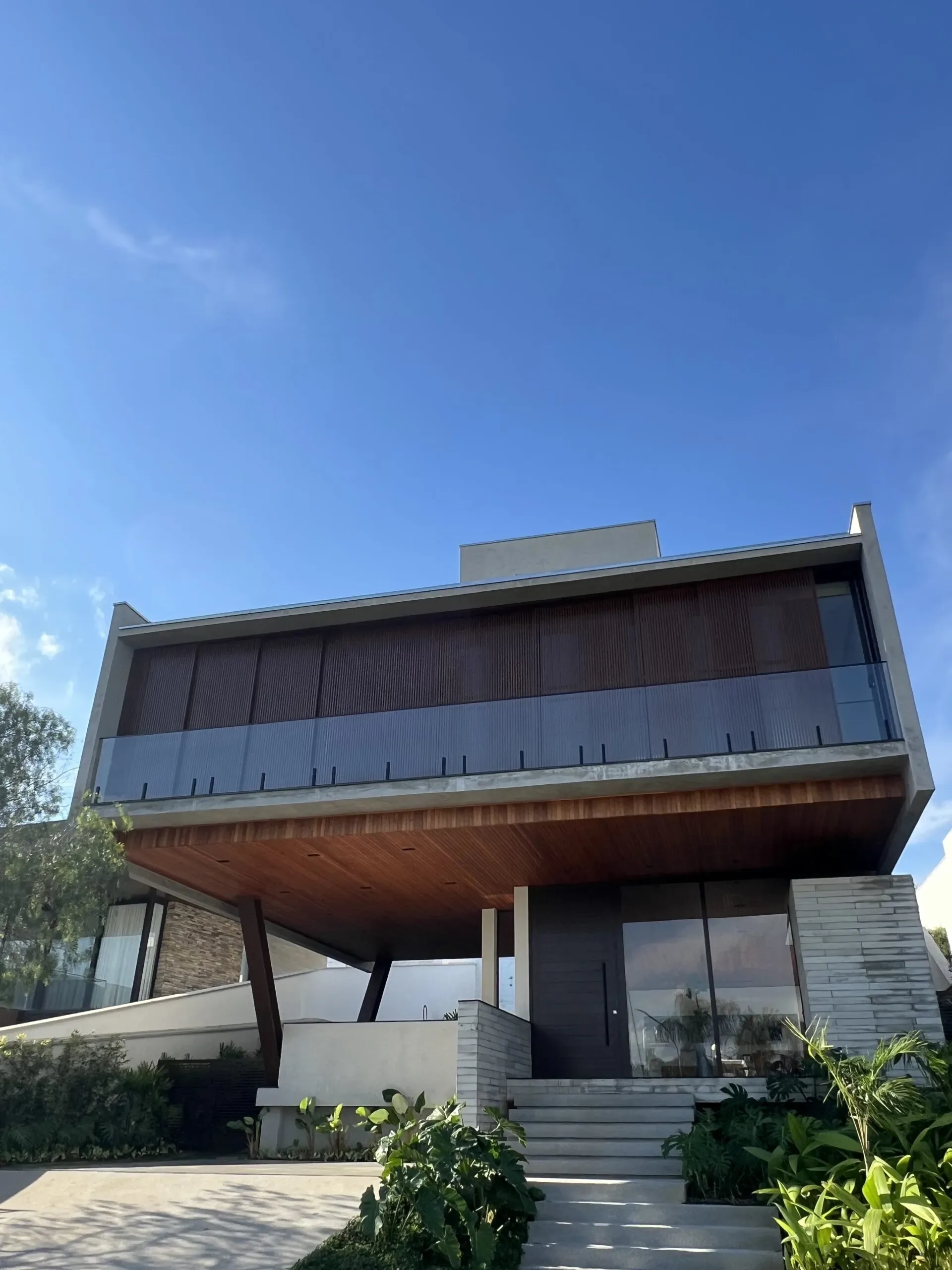 Vista frontal em ângulo diferente da mesma residência de alto padrão, evidenciando o guarda-corpo de vidro com perfis pretos no segundo pavimento e a porta de entrada com esquadria escura.