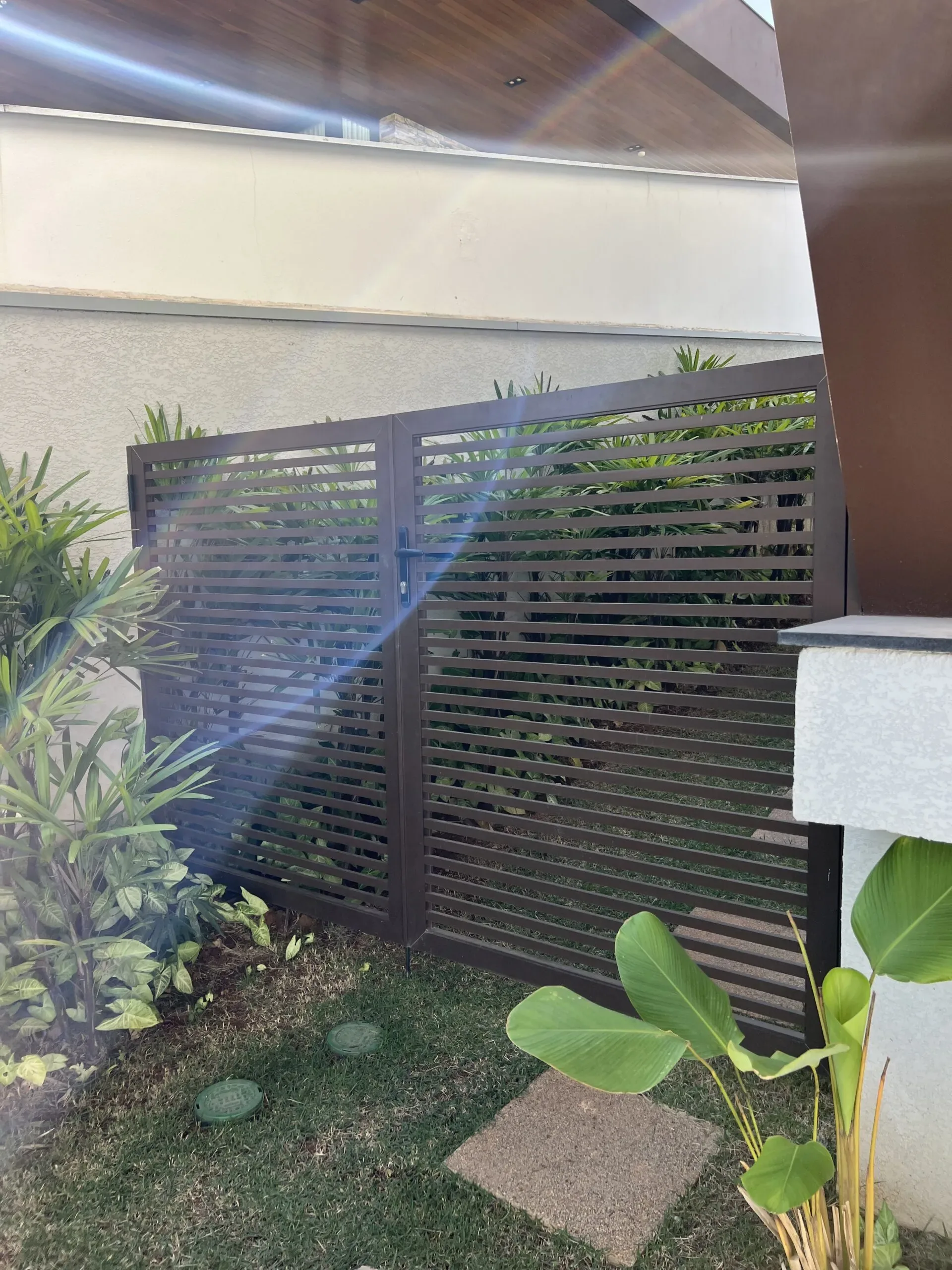 Portão duplo em alumínio ripado marrom, integrado a um corredor de paisagismo com plantas e gramado